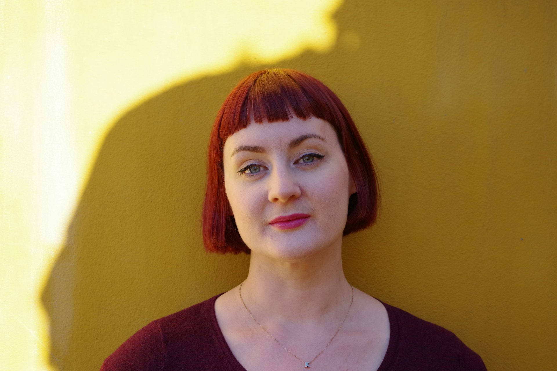 A white woman with a red short blunt cut bob and micro fringe leans against a yellow wall. She wears red lipstick,