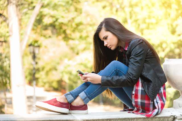 Wearing jeans and a black jacket while siting on a stone fence overlooking a leafy wooded area, a pensive teenage girl looks forlornly at her cell phone.