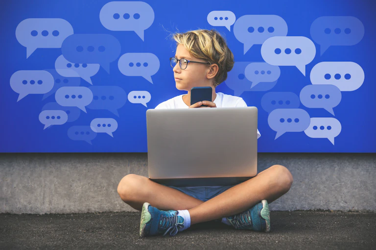 A boy with a laptop and holding a cell phone looks to the left. Conversation bubbles with dots are in the background.