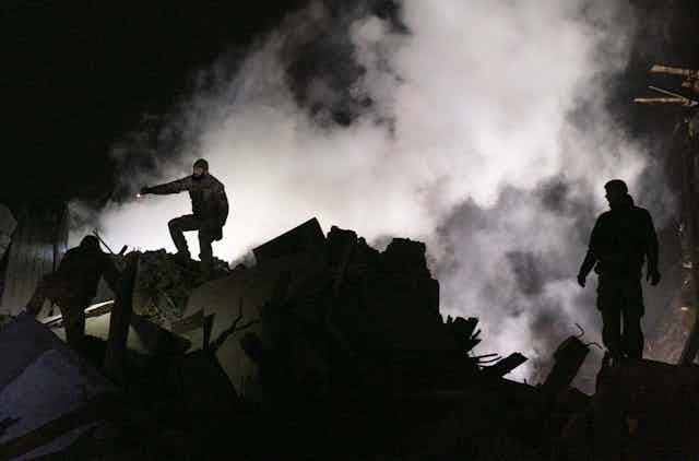Three Ukrainian rescue workers surrounded with smoke and silhouetted against the night sky.