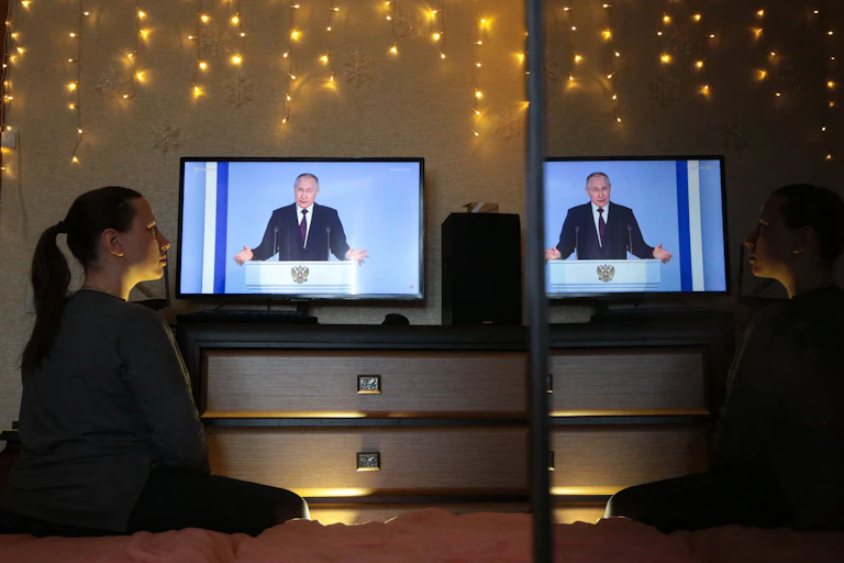 A woman looks to her right in a dark room with hanging lights, looking at a lit up television screen with a man in a suit speaking at a podium.