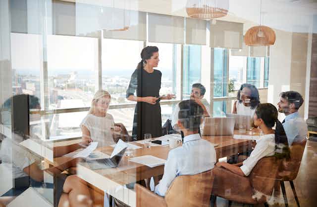 A woman leads a meeting around a conference room table in sunny office