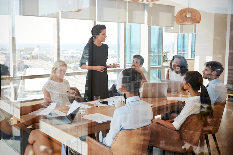 A woman leads a meeting around a conference room table in sunny office