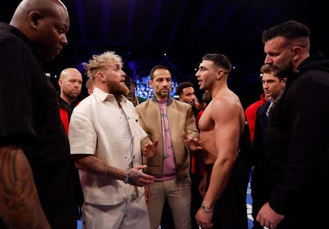 Jake Paul stands opposite Tommy Fury in a boxing ring. Paul has blonde hair and wears all white. Brunette Tommy is shirtless.