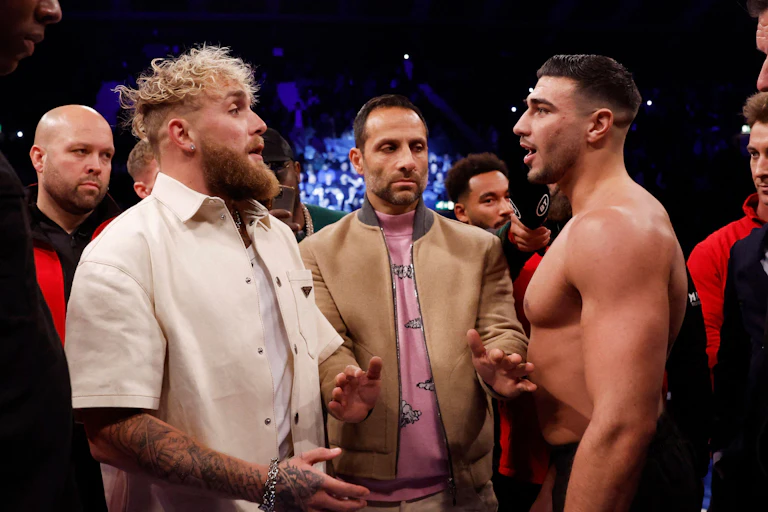 Jake Paul stands opposite Tommy Fury in a boxing ring. Paul has blonde hair and wears all white. Brunette Tommy is shirtless.