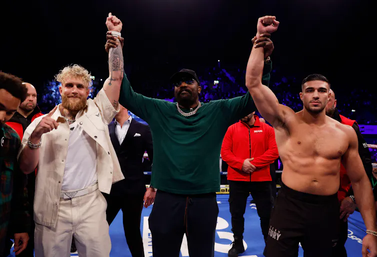 Dereck Chisora holds Fury and Paul's arms aloft in a boxing ring.