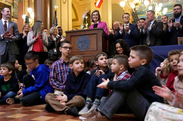 Children sit on a floor in front of adults clapping