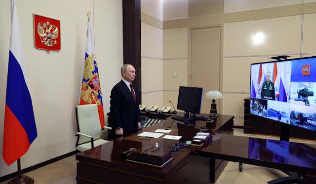Vladimir Putin stands behind his desk watching a new nuclear submarine being launched on a video screen.