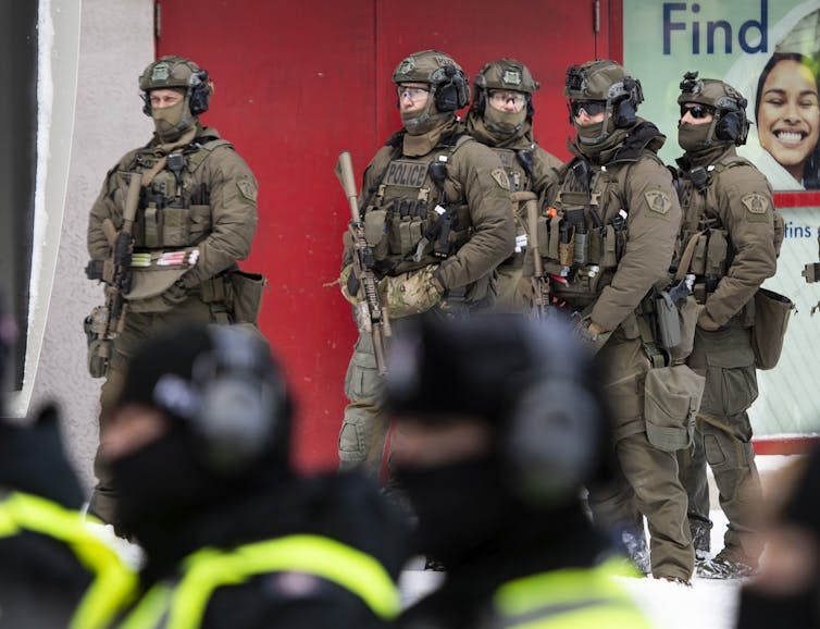 A group of RCMP officers in tactical gear stand behind a line of municipal police officers in fluorescent vests