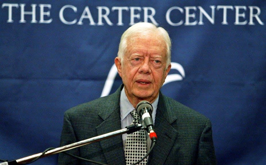 A gray-haired man stands and speaks into a microphone. Behind him, a blue banner with white writing reads: The Carter Center.