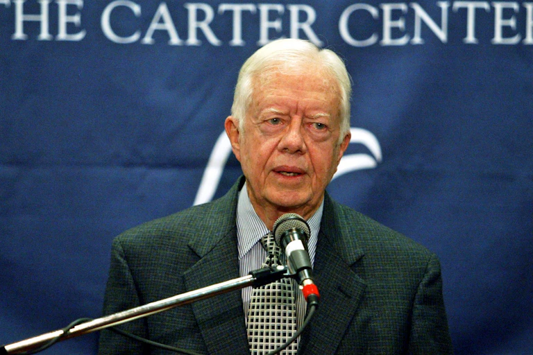 A gray-haired man stands and speaks into a microphone. Behind him, a blue banner with white writing reads: The Carter Center.