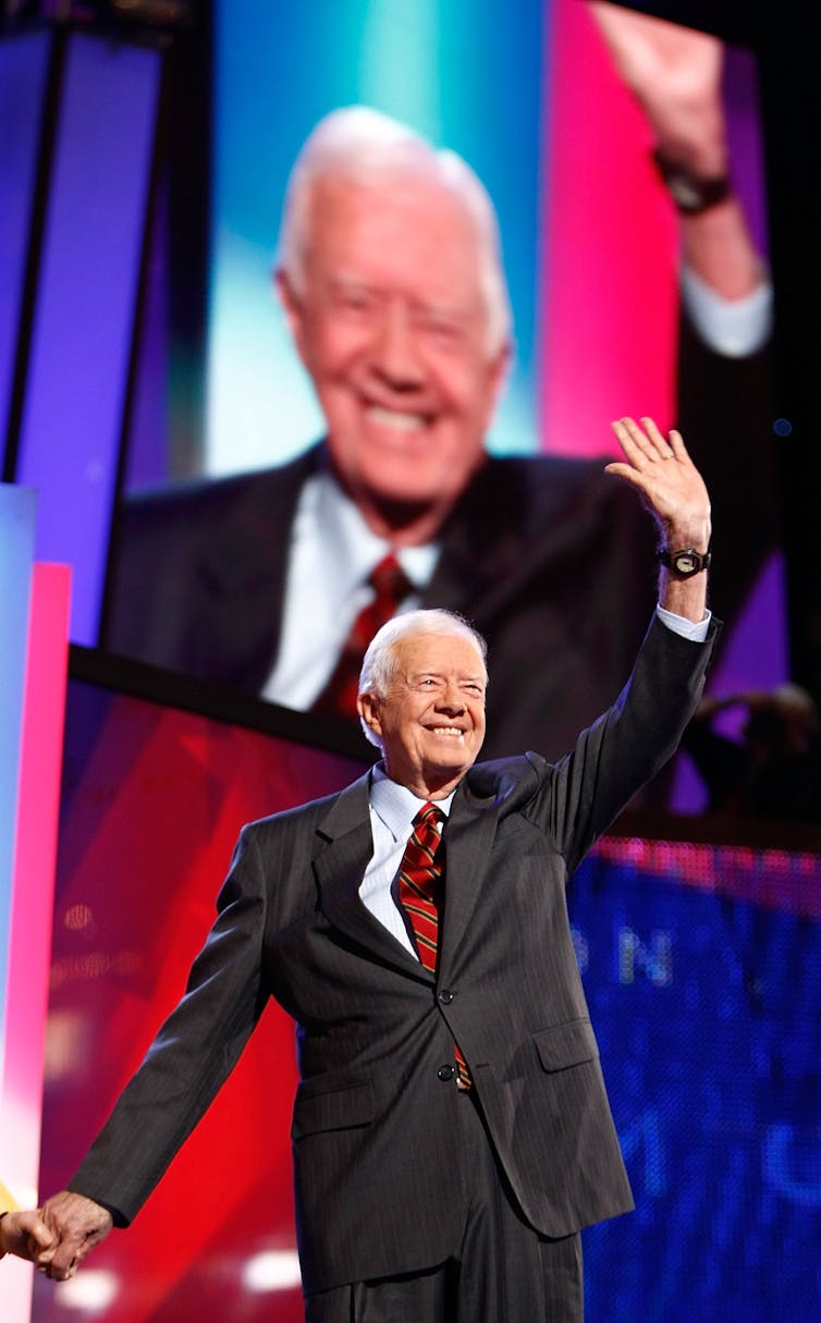 A suited, smiling gray-haired man walks on stage, with his left hand raised high, as he waves to the audience before him. Behind him, a large video screen captures his actions.