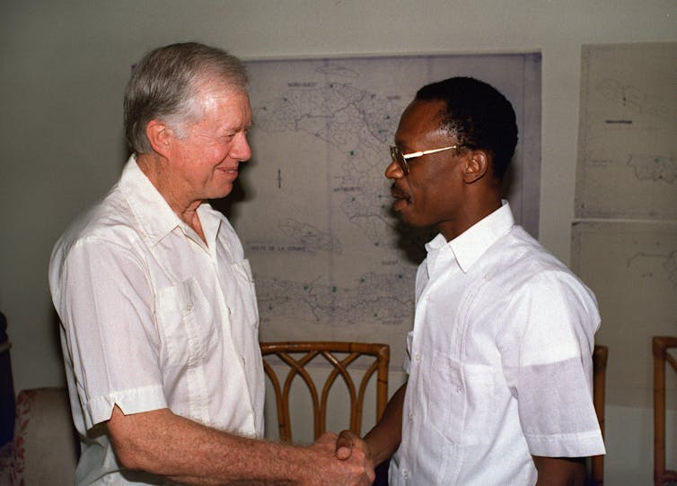 A gray-haired white man wearing a short-sleeved white shirt shakes the hand of a Black man, who is wearing glasses and a short-sleeved white shirt.