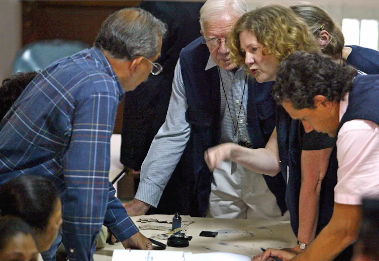 One man, on the left, stands leaning over a table as he speaks with a man, center, a woman and man on the right, who are also standing and leaning over the table.