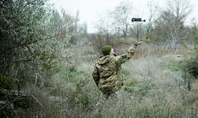 A small quadcopter hovers several feet above the outstretched hand of a soldier standing in a field