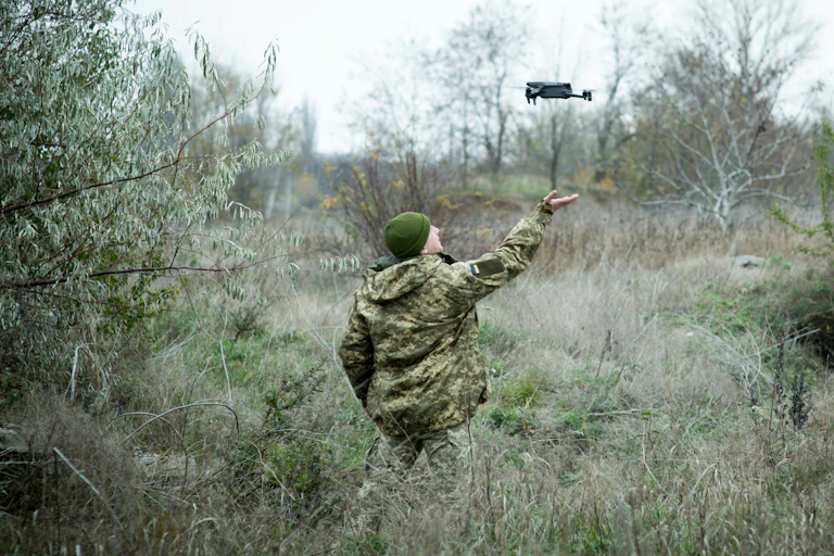 A small quadcopter hovers several feet above the outstretched hand of a soldier standing in a field