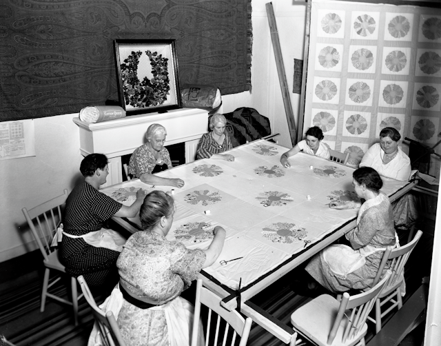 A black and white photo of a group of women sitting around a table sewing a quilt.
