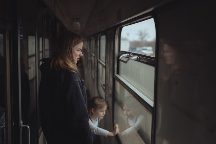 A young woman and her young daughter look out the window from a walkway aboard a train.