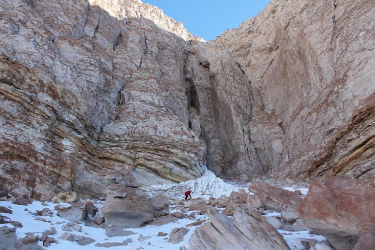 A human in a red coat looks tiny, crouching in front of enormous reddish rock formations that have ice in between.