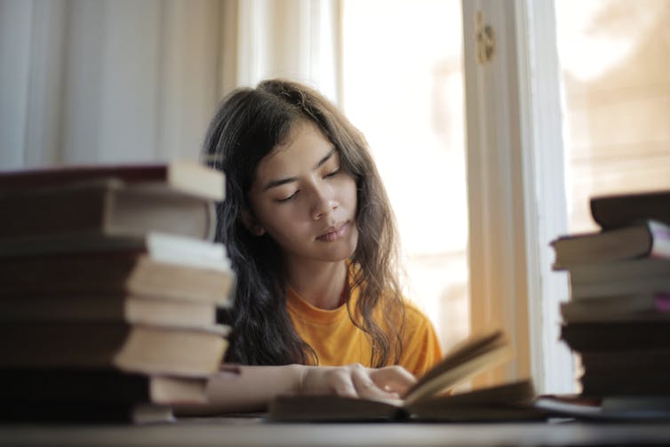 A young woman reads a book, with stacks of books next to her.