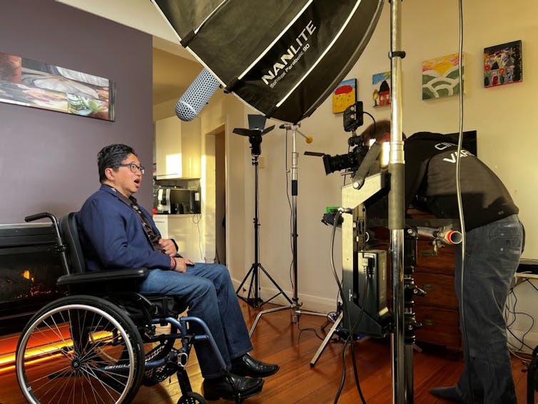 An Asian Australian man using a wheelchair, speaks directly to camera in a lounge room surrounded by lights, reflectors and microphones.