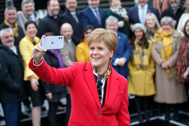 Nicola Sturgeon holds her iphone up to take a selfie, with other MSPs in a group behind her