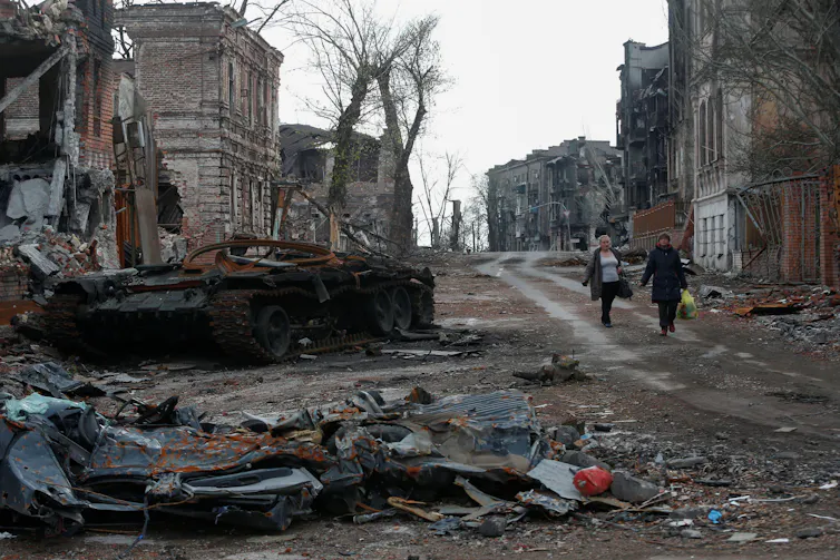 Two women walking past destroyed buildings.