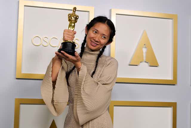 Chloe Zhao wears a beige dress with wide sleeves and holds up her Oscar in two hands. Her black hair is neatly parted in the middle and pulled into two plaits. 