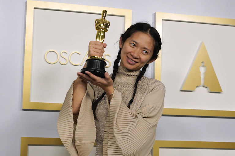 Chloe Zhao wears a beige dress with wide sleeves and holds up her Oscar in two hands. Her black hair is neatly parted in the middle and pulled into two plaits.