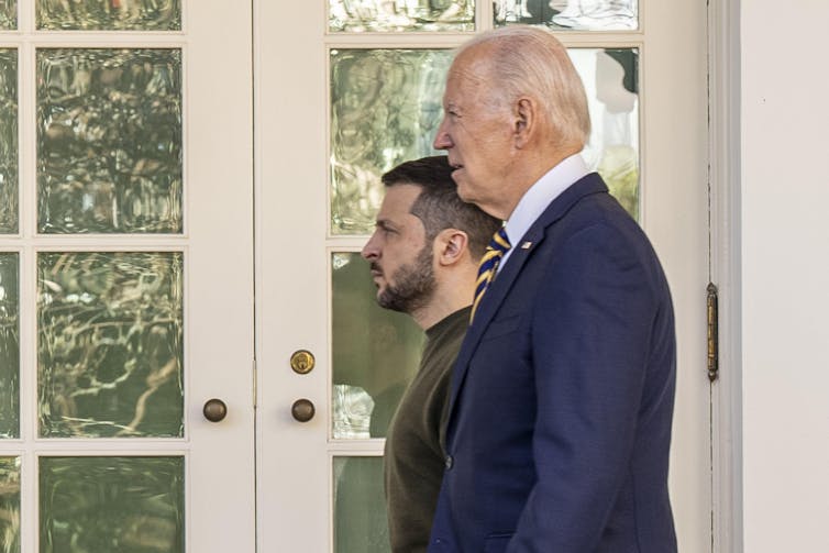 Two men walk past a white door.