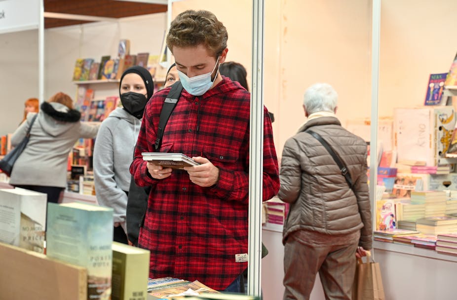 A young man in a mask reading a poetry book in a bookshop.