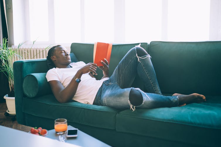 A young black man reclines on a sofa in a white t-shirt and jeans. He reads a red book.
