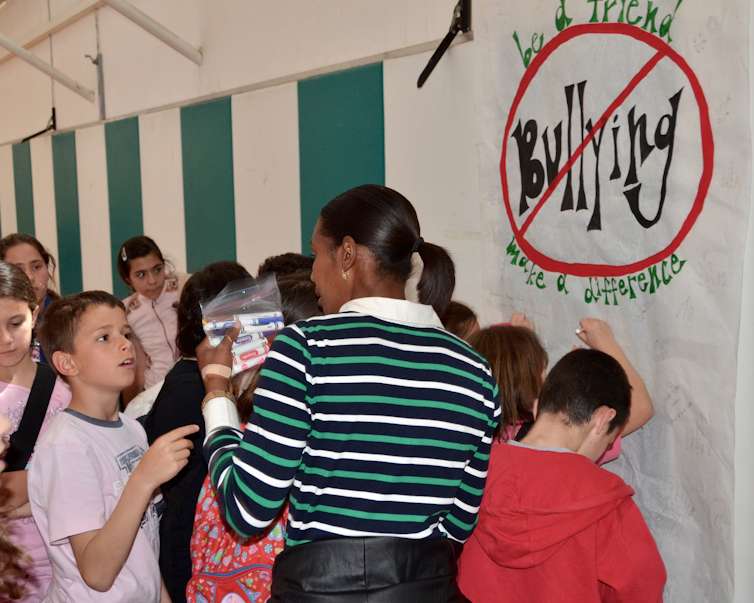 Kids and a teacher in the hallway of a school making a no bullying sign.