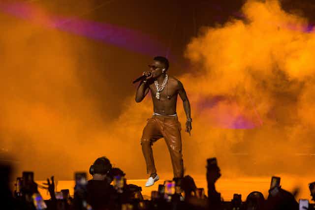 Wizkid performs shirtless in leather brown trousers against a backdrop of orange smoke.
