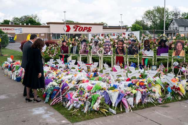 Two people wearing dark clothing look over a large bunch of flowers with photos of people, in front of a store that says Tops.