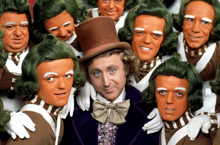 Gene Wilder wears a top hat, surrounded by orange-faced Oompa-Loompas.