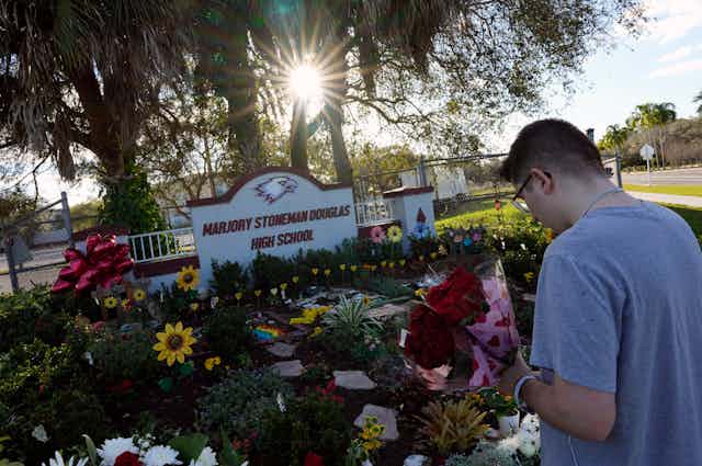 A person holds flowers in front of a pile of flowers and a sign reading 'Marjory Stoneman Douglas High School'