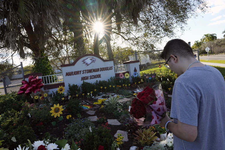 A person holds flowers in front of a pile of flowers and a sign reading 'Marjory Stoneman Douglas High School'