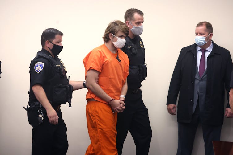 A white man in an orange prison suit wears a white face mask and has his hands folded together. Two police officers walk on either side of him, holding his arms.