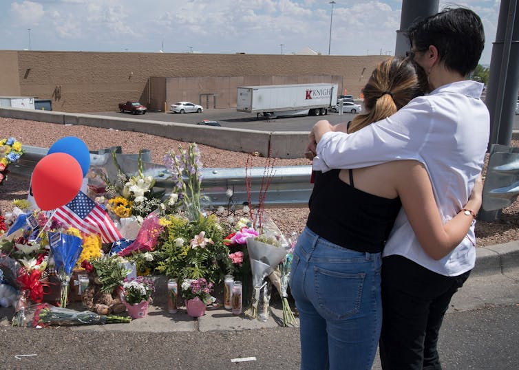 Two people face towards a large red brick building and a small pile of flowers, balloons and an American flag.