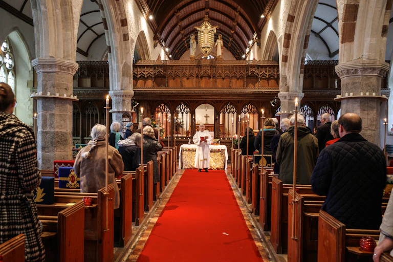 People praying inside a church with a priest in a white robe standing in front of the pews leading the prayers.