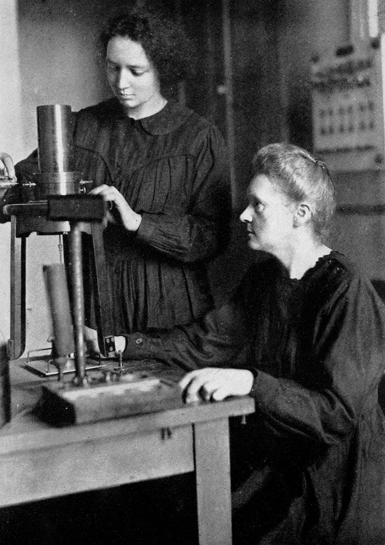 Czarno-białe zdjęcie Marie Curie siedzącej przed biurkiem i obserwującej, jak jej córka dostosowuje instrument.