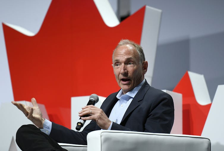 A middle-aged man in a suit sits in an arm chair speaking into a microphone