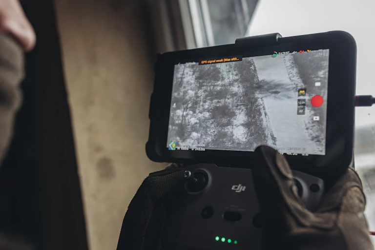 a pair of hands hold a small flight controller with an attached smart phone, the screen showing an aerial view of a winter landscape