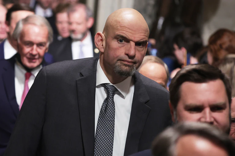 Wearing a black jacket, white shirt and checkered tie, U.S. Senator John Fetterman walks down a corridor in the U.S. Capitol along with his Congressional colleagues.