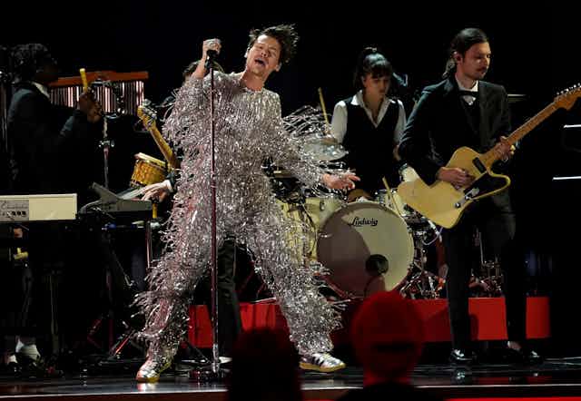 Harry Styles performing on stage wearing a metallic silver fringed onesie by Gucci