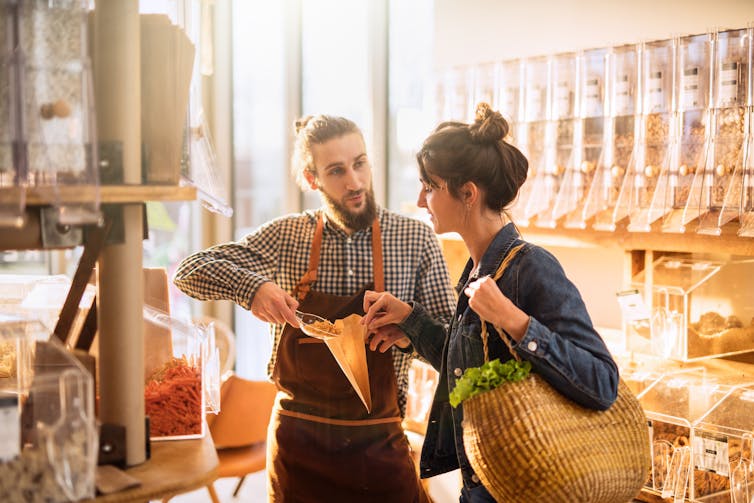 Young woman shopping in a light, bright bulk food store, being shown what to do by worker.