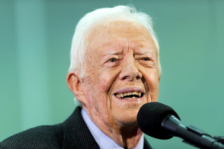 Former US President Jimmy Carter in a suit, speaking into a microphone