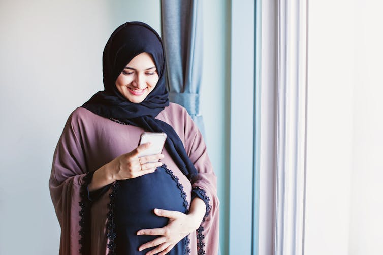 Pregnant Muslim woman clutching belly looking at phone in hand in front of window