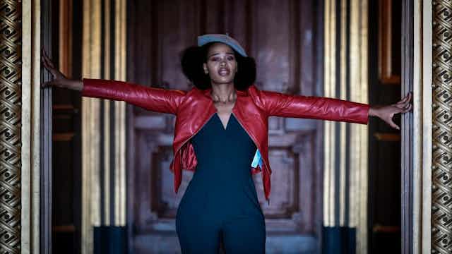 A woman with an Afro coming out from beneath a beret poses with her arms out in a gilded doorway, wearing a red leather jacket and a no-nonsense expression.
